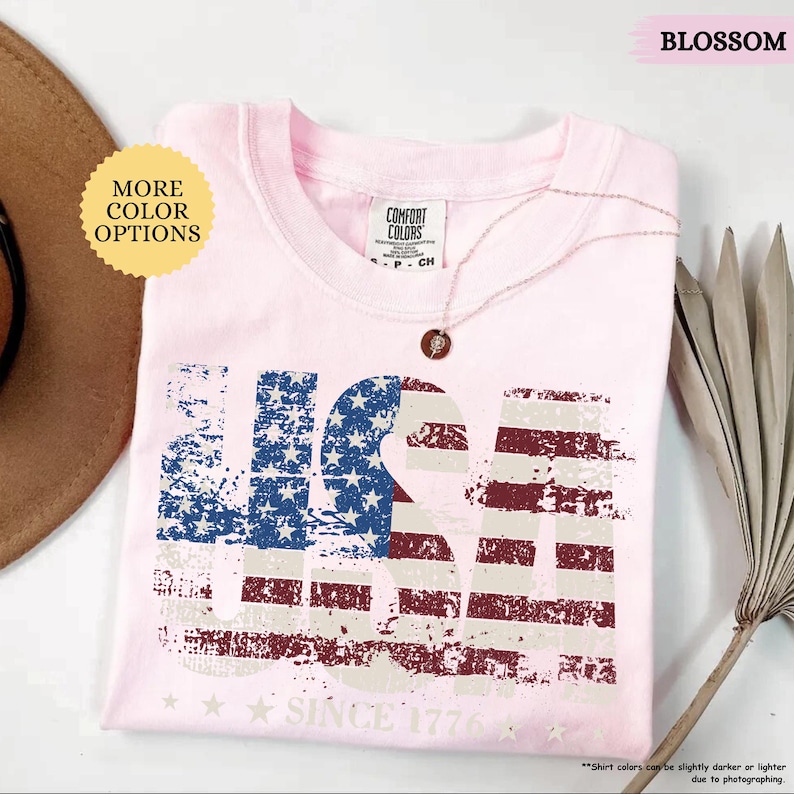 May include: A light pink t-shirt with a distressed USA graphic in blue, white, and red. The text "SINCE 1776" is below the graphic. The shirt is folded and has a Comfort Colors tag. A gold necklace with a pendant is on the shirt.