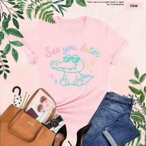 May include: A pink t-shirt with the phrase "See you later" and a cartoon alligator wearing sunglasses. The shirt is paired with blue jeans, a brown handbag, and black heels. Green leaves and foliage surround the items.