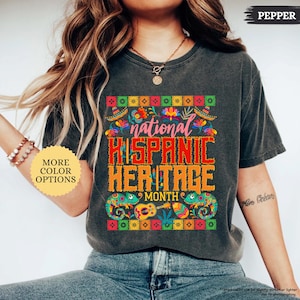 Peut inclure: T-shirt gris fonc&eacute; avec l'inscription "national HISPANIC HERITAGE MONTH" en rouge et orange. Le motif comprend des motifs floraux et g&eacute;om&eacute;triques color&eacute;s, et deux cam&eacute;l&eacute;ons. Le t-shirt porte &eacute;galement le mot "PEPPER".