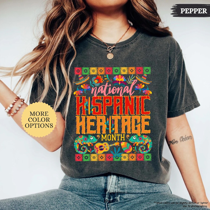 Peut inclure: T-shirt gris fonc&eacute; avec l'inscription "national HISPANIC HERITAGE MONTH" en rouge et orange. Le motif comprend des motifs floraux et g&eacute;om&eacute;triques color&eacute;s, et deux cam&eacute;l&eacute;ons. Le t-shirt porte &eacute;galement le mot "PEPPER".