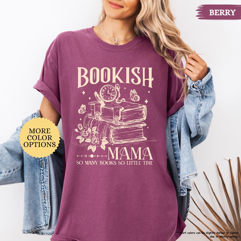 May include: Berry-coloured t-shirt with the text "BOOKISH MAMA" above a stack of books, a clock, and butterflies. The phrase "SO MANY BOOKS SO LITTLE TIME" is below the graphic. The shirt has rolled-up sleeves and a relaxed fit. "MORE COLOR OPTIONS" is in a yellow circle.