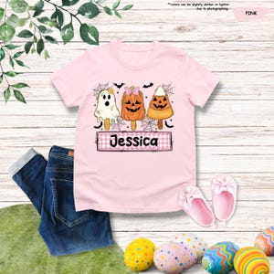 Może przedstawiać: Jasnor&oacute;żowy t-shirt z grafiką przedstawiającą trzy lody na patyku w stylu Halloween: białego duszka, pomarańczową dynię i cukierek kukurydziany. Poniżej znajduje się r&oacute;żowy prostokąt w kratkę z nadrukowanym czarnym napisem Jessica.