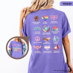 May include: A violet-colored t-shirt with various graphic designs and text promoting social justice and equality. The shirt includes phrases like "Black Lives Matter," "Love is Love," and "Feminism is for Everyone."