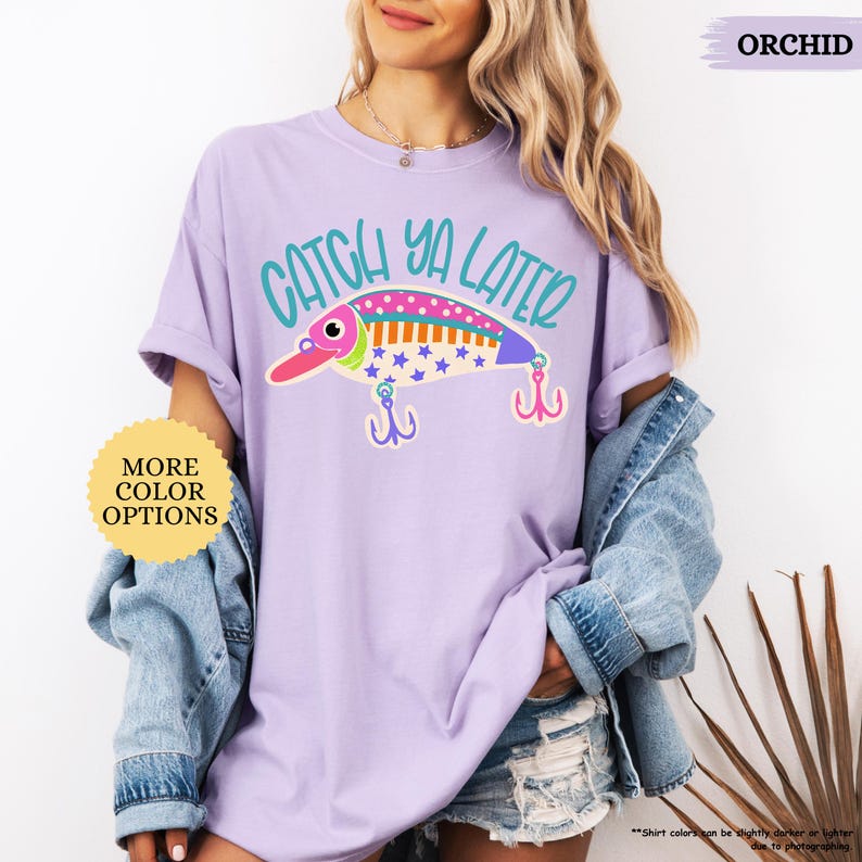 以下が含まれることがあります： オーキッドカラーのTシャツで、カラフルな釣り用ルアーのグラフィックとティールカラーの「CATCH YA LATER」という文字がプリントされています。ルアーのデザインにはピンク、オレンジ、ブルーのストライプと青い星があしらわれ、下に2つのピンクのトリプルフックが描かれています。