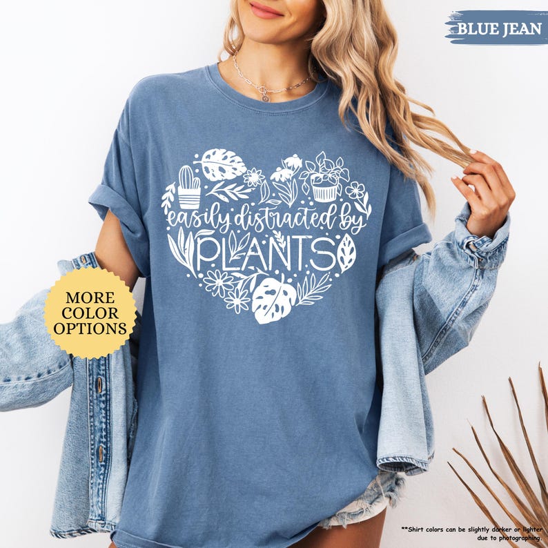 Pu&ograve; includere: Una t-shirt blu jeans con un design a forma di cuore bianco con il testo "easily distracted by PLANTS" e una variet&agrave; di illustrazioni di piante. La t-shirt &egrave; mostrata su un manichino che indossa una giacca di jeans.