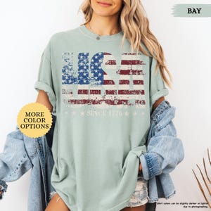 May include: A light green t-shirt with a distressed USA flag design in red, white, and blue. The text "SINCE 1776" and stars are below the flag. The shirt has rolled-up sleeves and is part of a collection with more color options.