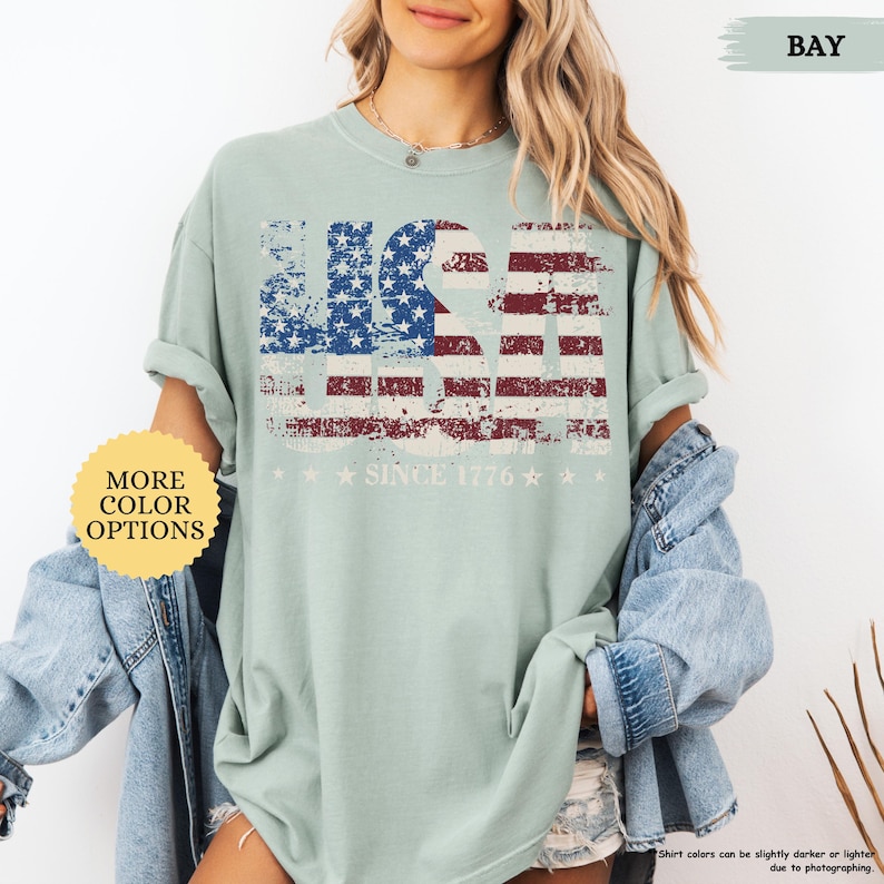May include: A light green t-shirt with a distressed USA flag design in red, white, and blue. The text "SINCE 1776" and stars are below the flag. The shirt has rolled-up sleeves and is part of a collection with more color options.