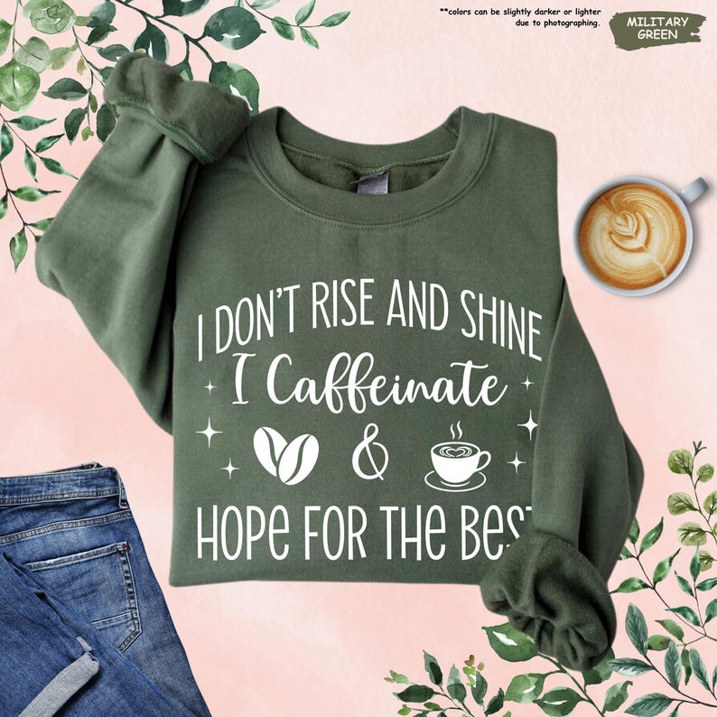 May include: Military green sweatshirt with the text "I DON'T RISE AND SHINE I Caffeinate & HOPE FOR THE BEST" in white. The design includes coffee cup and coffee bean graphics. A cup of latte art is in the upper right corner.