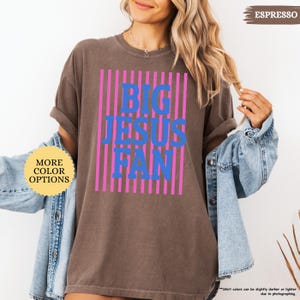 May include: Espresso-colored t-shirt with the text "BIG JESUS FAN" in blue, set against a background of pink vertical stripes. The shirt has rolled-up sleeves and is worn with a denim jacket. The words "MORE COLOR OPTIONS" are in a yellow circle.