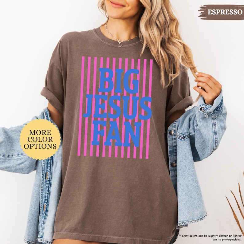 May include: Espresso-colored t-shirt with the text "BIG JESUS FAN" in blue, set against a background of pink vertical stripes. The shirt has rolled-up sleeves and is worn with a denim jacket. The words "MORE COLOR OPTIONS" are in a yellow circle.
