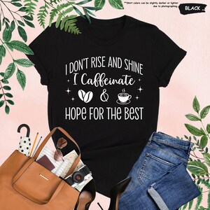 May include: Black t-shirt with the text "I DON'T RISE AND SHINE I Caffeinate & HOPE FOR THE BEST" in white. The design includes coffee beans and a coffee cup. The shirt is paired with blue jeans and a brown tote bag.