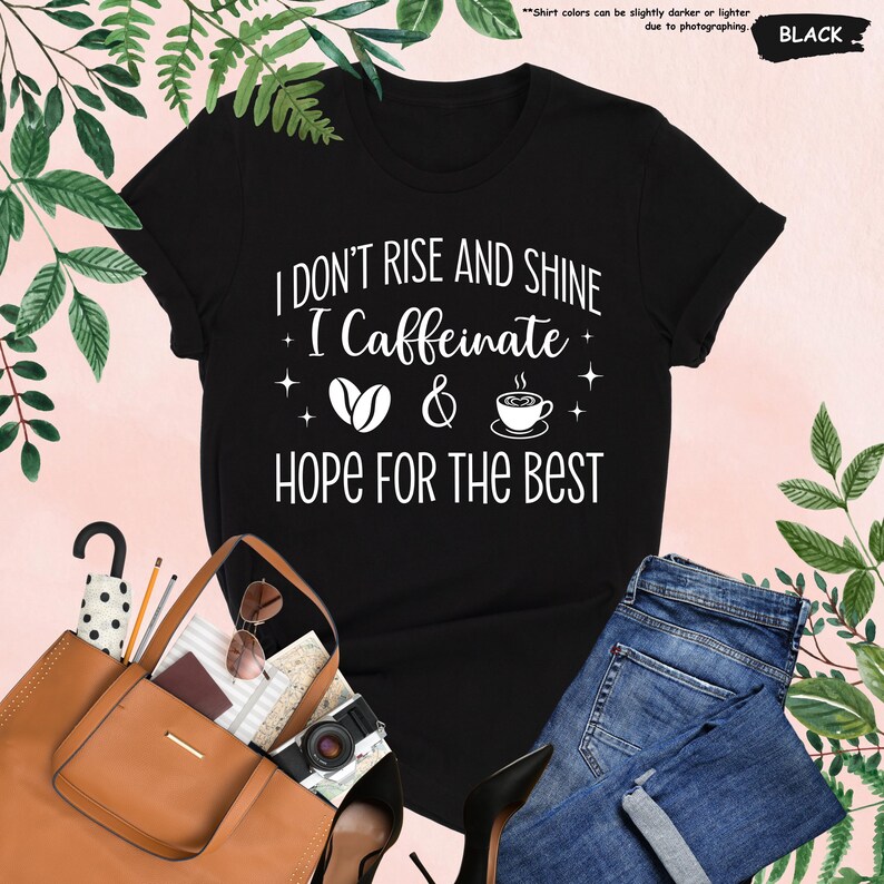 May include: Black t-shirt with the text "I DON'T RISE AND SHINE I Caffeinate & HOPE FOR THE BEST" in white. The design includes coffee beans and a coffee cup. The shirt is paired with blue jeans and a brown tote bag.