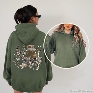 May include: A military green hoodie featuring a deer-themed graphic on the back, including deer, a hunting blind, and camouflage bows. The front of the hoodie has a small bow graphic. The hoodie has a hood and a front pocket.