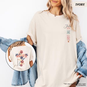 May include: A light beige t-shirt with the text "Jesus is the light" on the front and a floral cross design on the back. The shirt is shown on a model wearing a denim jacket.