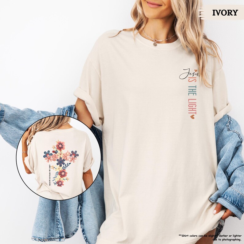 May include: A light beige t-shirt with the text "Jesus is the light" on the front and a floral cross design on the back. The shirt is shown on a model wearing a denim jacket.