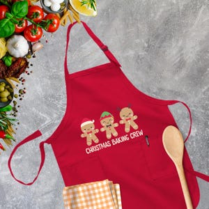 Op de afbeelding: Rode kerstbakschort met een zak en drie gingerbreadmen die hand in hand staan. De tekst op de schort staat "Christmas Baking Crew".