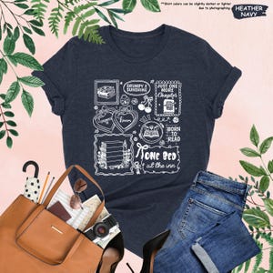May include: Heather navy t-shirt with a white graphic design featuring book-themed illustrations and text. The design includes phrases like "Grumpy x Sunshine," "Just One More Chapter," and "Born to Read." The shirt is paired with blue jeans and a brown tote bag.