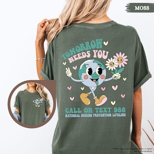 May include: Moss green t-shirt with a graphic on the back. The design shows a smiling Earth character holding flowers and giving a peace sign. Text reads "Tomorrow Needs You" and "Call or Text 988, National Suicide Prevention Lifeline."