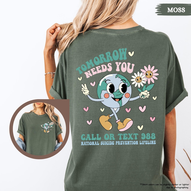 May include: Moss green t-shirt with a graphic on the back. The design shows a smiling Earth character holding flowers and giving a peace sign. Text reads "Tomorrow Needs You" and "Call or Text 988, National Suicide Prevention Lifeline."