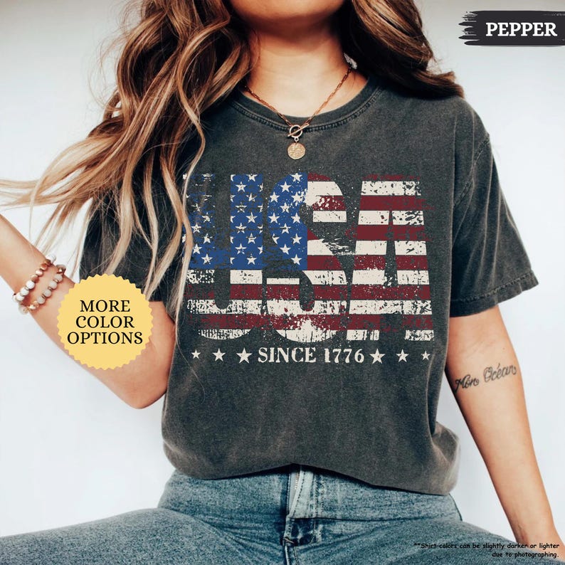 May include: A dark gray t-shirt with a distressed USA flag design. The flag features red and white stripes, blue stars, and the text "USA SINCE 1776". The shirt has a vintage, worn look.