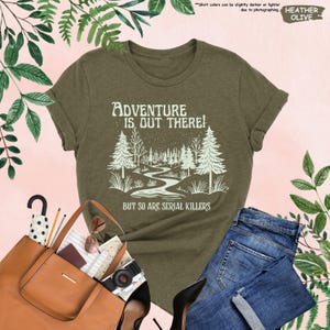 May include: Olive green t-shirt with the text "ADVENTURE IS OUT THERE! BUT SO ARE SERIAL KILLERS" and a graphic of a forest scene. The shirt is paired with blue jeans, a brown tote bag, and accessories.
