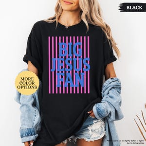May include: Black t-shirt with the text "BIG JESUS FAN" in blue letters, set against a background of pink vertical stripes. The shirt has short sleeves and a round neckline. A button with the text "MORE COLOR OPTIONS" is visible.