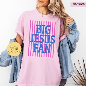 May include: A light pink t-shirt with the text "BIG JESUS FAN" in blue, set against a background of pink vertical stripes. The shirt also has the text "MORE COLOR OPTIONS" in a yellow circle.