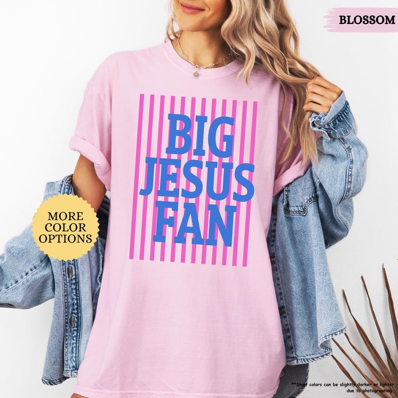 May include: A light pink t-shirt with the text "BIG JESUS FAN" in blue, set against a background of pink vertical stripes. The shirt also has the text "MORE COLOR OPTIONS" in a yellow circle.
