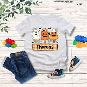 Może przedstawiać: Biały t-shirt z kr&oacute;tkim rękawem z grafiką przedstawiającą trzy lody na patyku w stylu Halloween: białego duszka, pomarańczową dynię w czapce oraz cukierek kukurydziany. Poniżej znajduje się pomarańczowa etykieta w kratkę z imieniem Thomas.