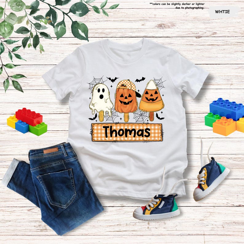 Może przedstawiać: Biały t-shirt z kr&oacute;tkim rękawem z grafiką przedstawiającą trzy lody na patyku w stylu Halloween: białego duszka, pomarańczową dynię w czapce oraz cukierek kukurydziany. Poniżej znajduje się pomarańczowa etykieta w kratkę z imieniem Thomas.