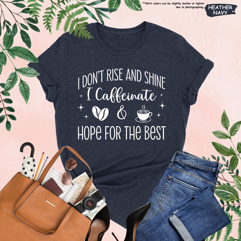 May include: Heather navy blue t-shirt with the text "I DON'T RISE AND SHINE I Caffeinate & HOPE FOR THE BEST". The shirt features coffee bean and coffee cup graphics. The shirt is paired with blue jeans and a brown tote bag.