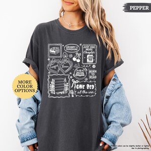 Puede incluir: Camiseta gris oscuro con dise&ntilde;os gr&aacute;ficos blancos, incluyendo libros, corazones y texto. El texto incluye frases como "Grumpy x Sunshine", "Just One More Chapter", "Born to Read" y "One Bed at the Inn".