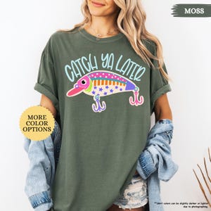 以下が含まれることがあります： モスグリーンの半袖Tシャツ。ライトブルーの文字で「CATCH YA LATER」と書かれ、その下にカラフルな釣り用ルアーのグラフィックが描かれています。ルアーはピンク、白、青で星柄があり、2つのピンクのトリプルフックが付いています。