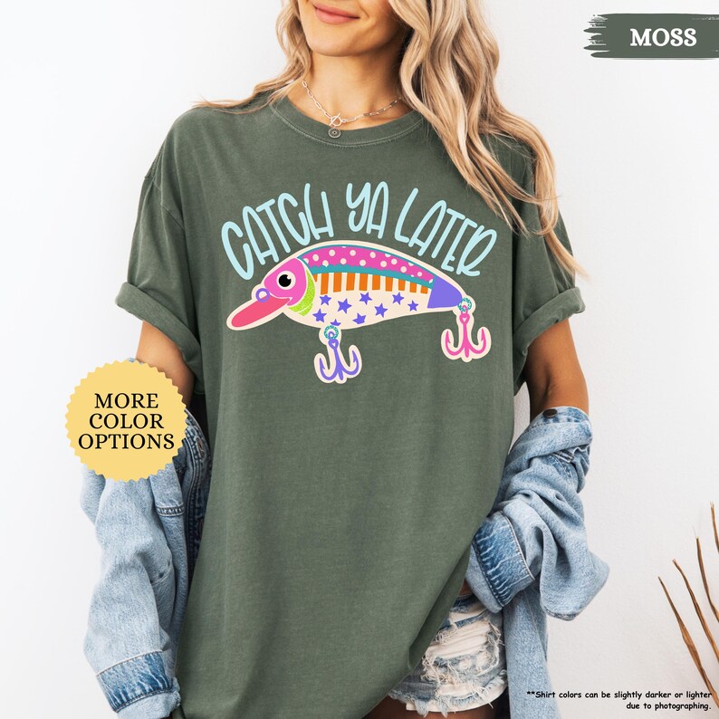 以下が含まれることがあります： モスグリーンの半袖Tシャツ。ライトブルーの文字で「CATCH YA LATER」と書かれ、その下にカラフルな釣り用ルアーのグラフィックが描かれています。ルアーはピンク、白、青で星柄があり、2つのピンクのトリプルフックが付いています。