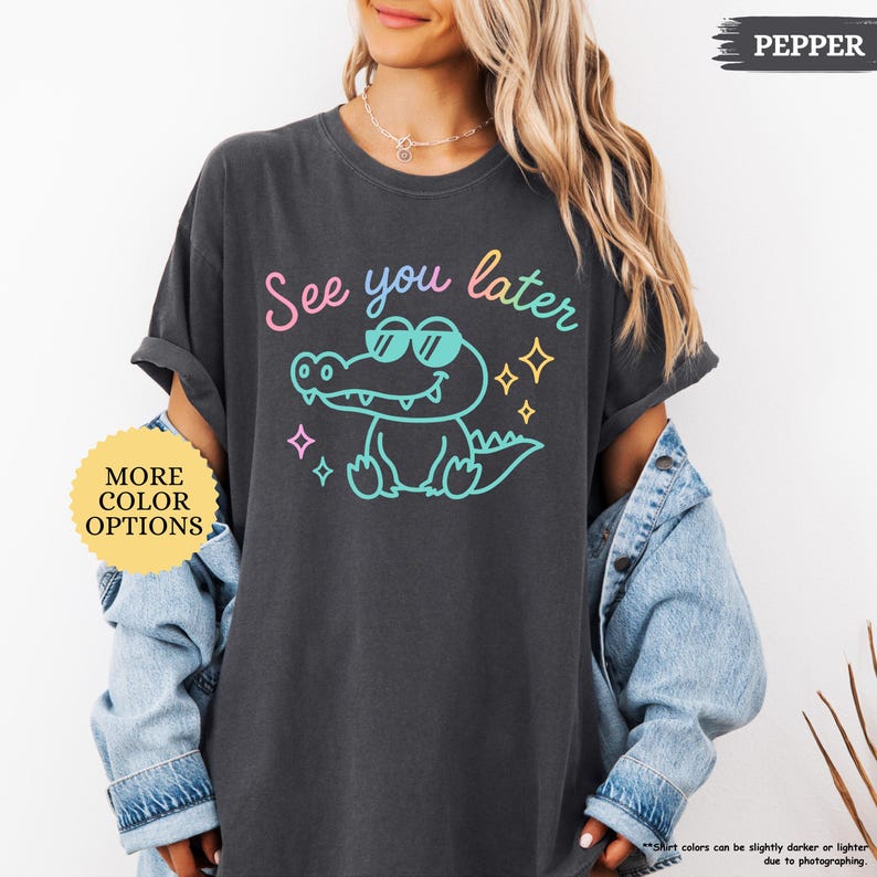 Puede incluir: Camiseta gris oscuro con la frase "See you later" sobre un cocodrilo de dibujos animados con gafas de sol. El cocodrilo est&aacute; delineado en turquesa, con acentos rosas, amarillos y turquesas. Un c&iacute;rculo amarillo con "MORE COLOR OPTIONS" est&aacute; en la parte inferior izquierda.