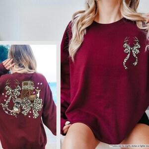 May include: Maroon-colored sweatshirt featuring a deer antler bow design on the front and a larger graphic on the back. The back graphic includes deer, a hunting blind, and a bow with a leopard print pattern. The sweatshirt is a long-sleeved crew neck.
