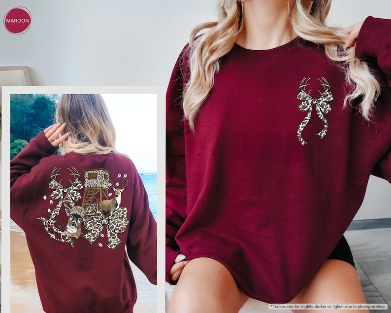 May include: Maroon-colored sweatshirt featuring a deer antler bow design on the front and a larger graphic on the back. The back graphic includes deer, a hunting blind, and a bow with a leopard print pattern. The sweatshirt is a long-sleeved crew neck.