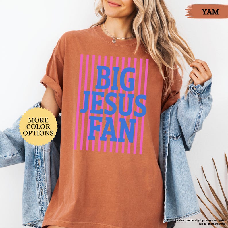 May include: A terracotta-colored t-shirt with the text "BIG JESUS FAN" in blue letters, set against a background of pink vertical stripes. The shirt has rolled-up sleeves and a casual fit. The words "MORE COLOR OPTIONS" are in a yellow circle.