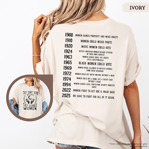 May include: An ivory t-shirt featuring a timeline of historical milestones in black text on the back, listing dates from 1900 to 2025. An inset circle shows the front of the shirt with a graphic of hands, a moon, and the text They didn't burn witches, they burned women.
