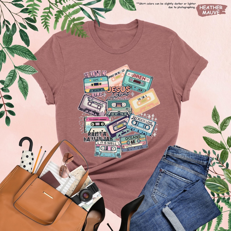 May include: Heather mauve t-shirt featuring a graphic of vintage cassette tapes with the words "Jesus Anthems" and other song titles. The design includes various colors and fonts. The shirt is paired with blue jeans and accessories.
