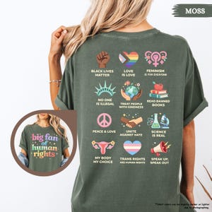 May include: Moss green t-shirt with a graphic of various social justice symbols and phrases, including "Black Lives Matter," a rainbow heart, and "Feminism is for Everyone." The shirt also features the text "big fan of human rights."