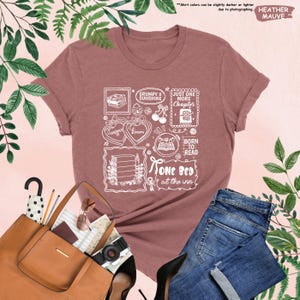 May include: Heather mauve t-shirt with a white graphic design featuring book-themed illustrations and text. The design includes the phrases "Grumpy x Sunshine", "Just One More Chapter", "Born to Read", and "One Bed at the Inn".