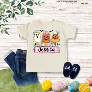 Może przedstawiać: T-shirt w naturalnym kolorze z grafiką przedstawiającą trzy lody na patyku w stylu Halloween: białego duszka, pomarańczową dynię z r&oacute;żową kokardką oraz cukierek kukurydziany. Poniżej znajduje się r&oacute;żowy prostokąt w kratkę z imieniem Jessica.