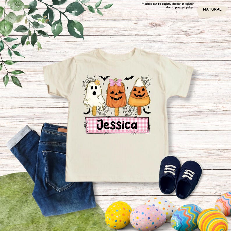 Może przedstawiać: T-shirt w naturalnym kolorze z grafiką przedstawiającą trzy lody na patyku w stylu Halloween: białego duszka, pomarańczową dynię z r&oacute;żową kokardką oraz cukierek kukurydziany. Poniżej znajduje się r&oacute;żowy prostokąt w kratkę z imieniem Jessica.