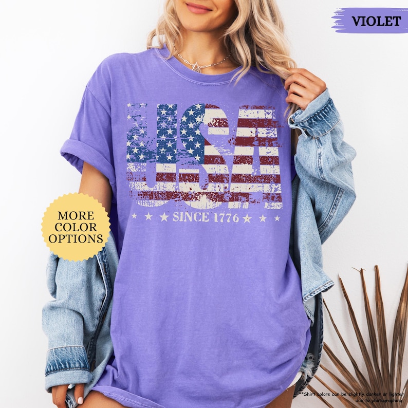 May include: A violet-colored t-shirt with a distressed USA flag design. The flag is incorporated into the word "USA" with stars and stripes. The text "SINCE 1776" is below the word "USA". The shirt has a round neck and short sleeves.