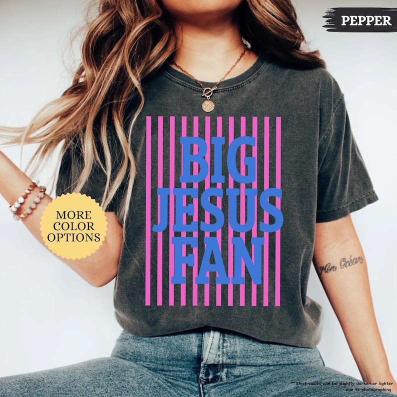 May include: Dark gray t-shirt with the text "BIG JESUS FAN" in blue letters, set against a pink striped background. The shirt has a vintage wash look and a crew neck. The words "MORE COLOR OPTIONS" are in a yellow circle.