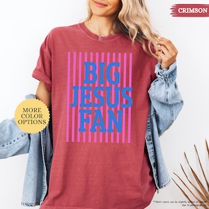 May include: Crimson-colored t-shirt with the text "BIG JESUS FAN" in blue letters, set against a background of pink vertical stripes. The shirt has a crew neck and short sleeves. The words "MORE COLOR OPTIONS" are in a yellow circle.