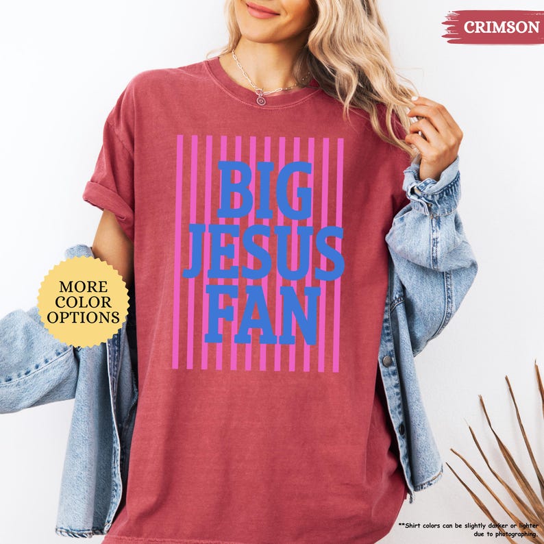 May include: Crimson-colored t-shirt with the text "BIG JESUS FAN" in blue letters, set against a background of pink vertical stripes. The shirt has a crew neck and short sleeves. The words "MORE COLOR OPTIONS" are in a yellow circle.