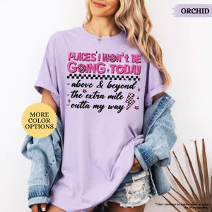 May include: Orchid-colored t-shirt with the text "PLACES I WON'T BE GOING TODAY" in pink and black. The shirt features additional text and graphics, including a checkered flag and lightning bolt. A round sticker on the left says "MORE COLOR OPTIONS".