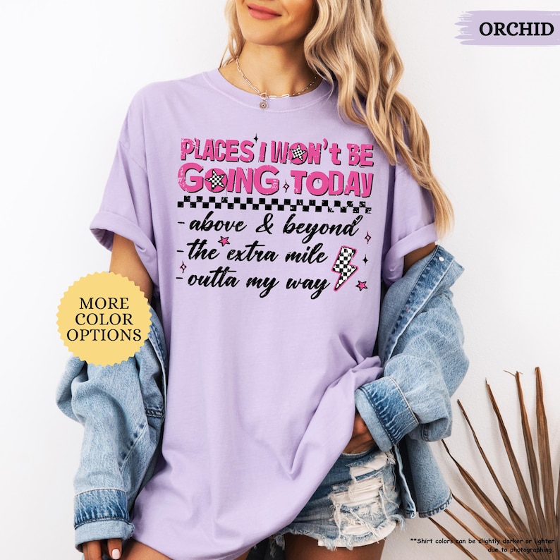 May include: Orchid-colored t-shirt with the text "PLACES I WON'T BE GOING TODAY" in pink and black. The shirt features additional text and graphics, including a checkered flag and lightning bolt. A round sticker on the left says "MORE COLOR OPTIONS".